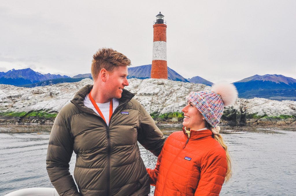 man and woman look at eachother in front of the lighthouse at the end of the world, she wears fur pom hat