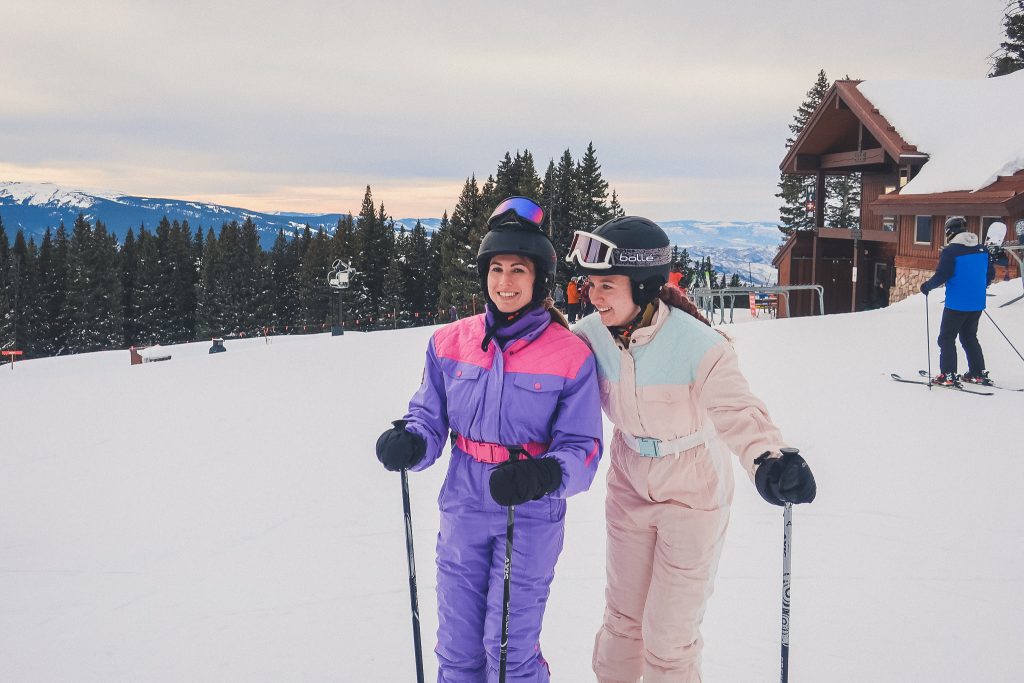 two women wear one piece snowsuits on the mountain in Vail, CO while skiing