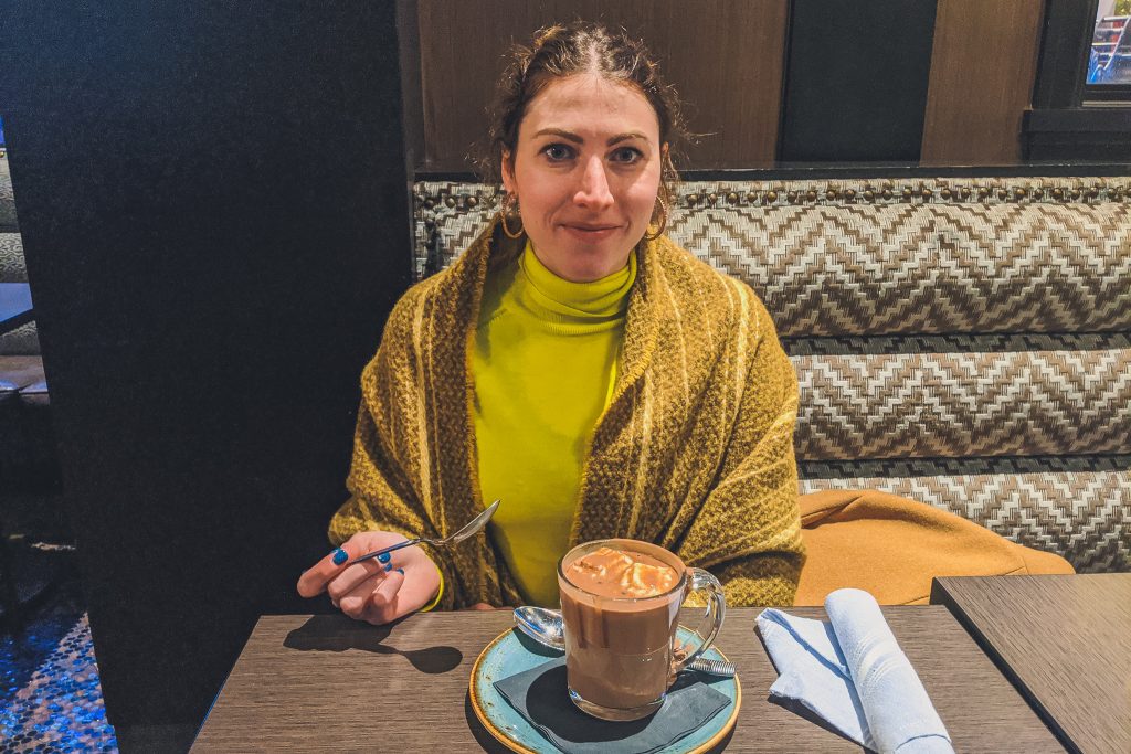 woman eats hot chocolate wearing long sleeve turtleneck and wool shawl 