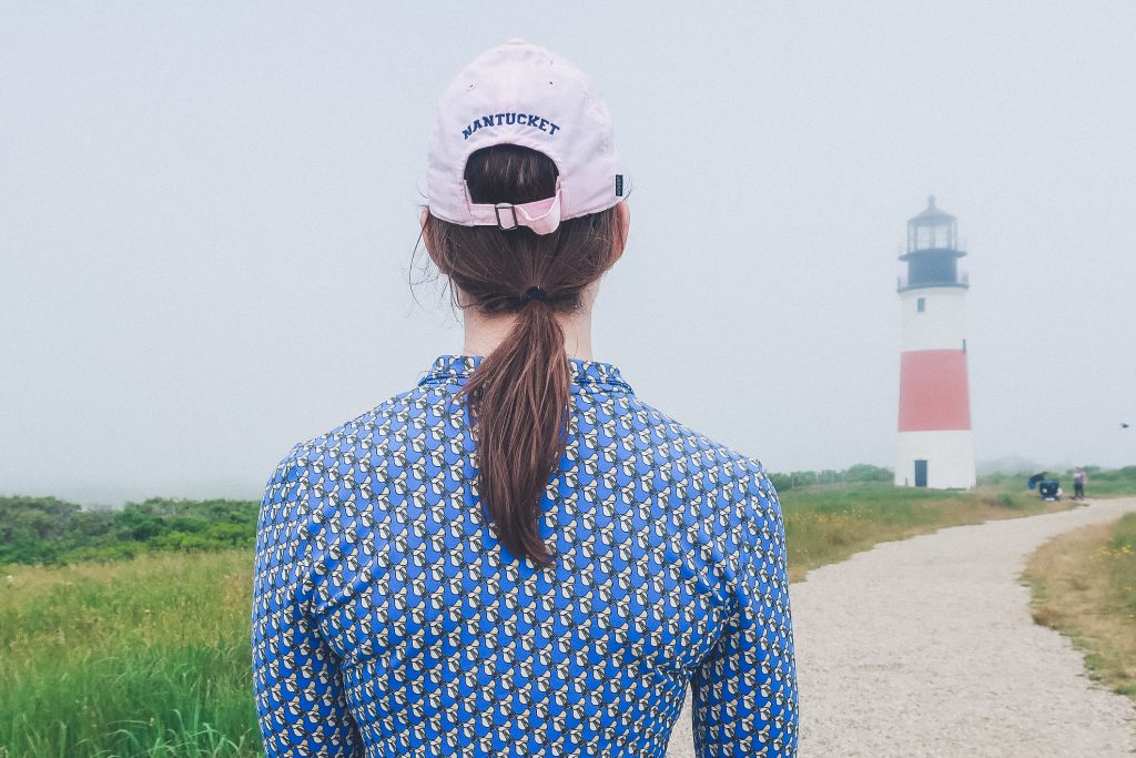 Sankaty Head lighthouse in the distance, woman looks out onto foggy day, wearing Nantucket logo ballcap
